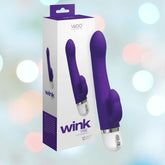 The image depicts an indigo VeDO Wink Rabbit Vibe, a personal massager intended for clitoral stimulation, positioned beside its packaging. The backdrop is gentle with softly blurred pastel lights. The box showcases the product image and the name "VeDO Wink Rabbit Vibe.